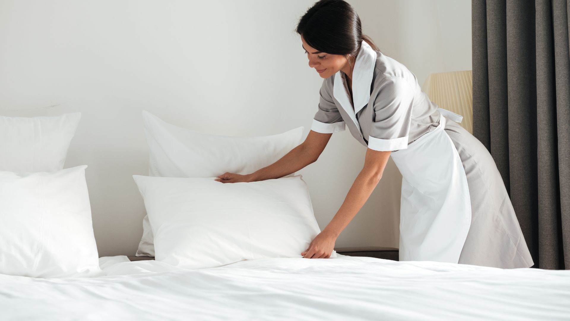 Housekeeper in uniform making a bed in a bright, clean hotel room