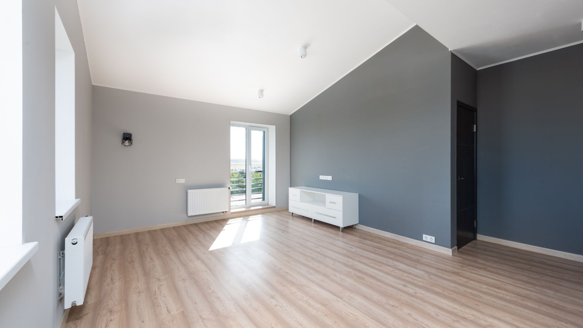 Empty room with gray walls, wooden floor, window, and white furniture