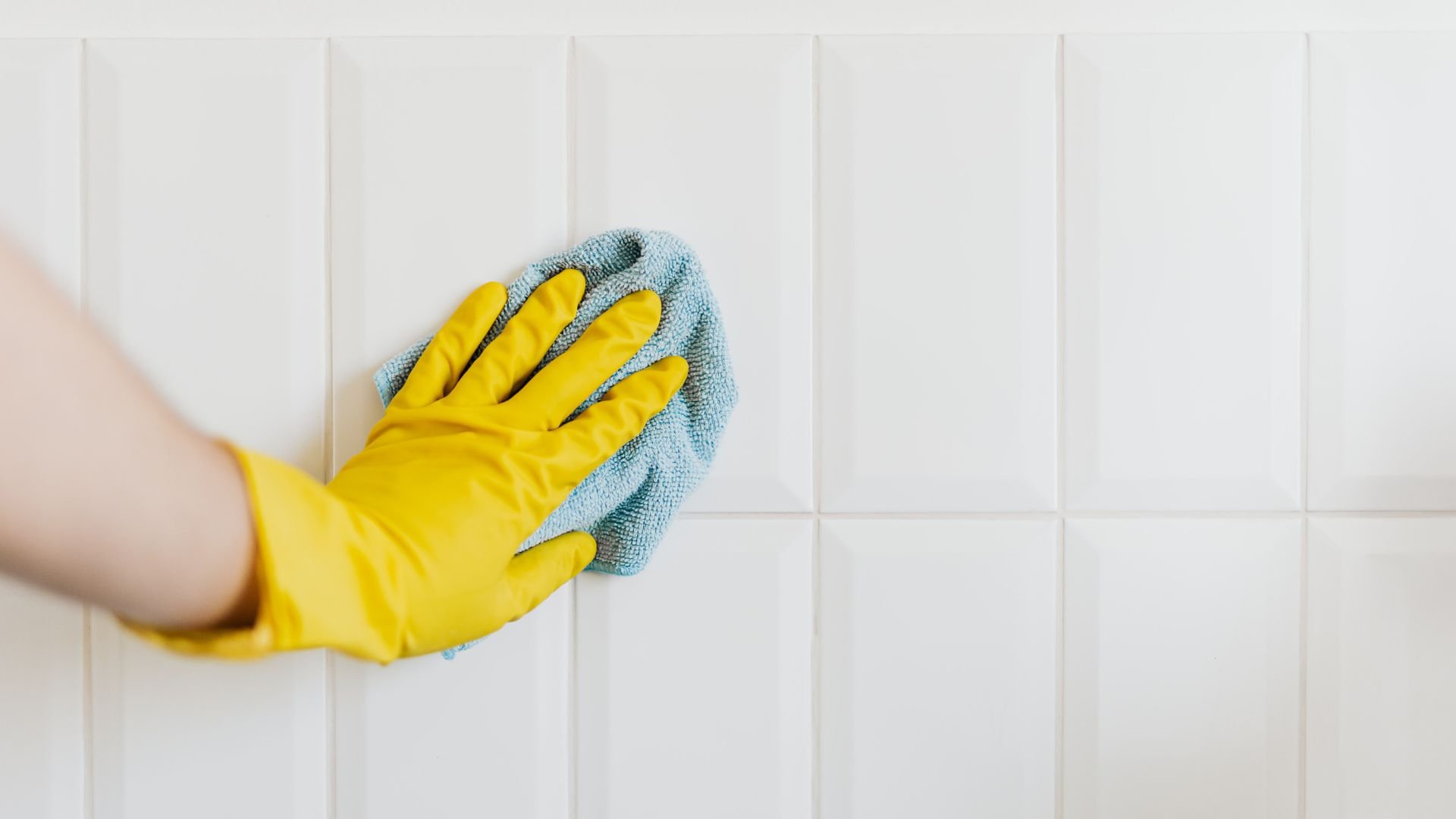 Yellow gloved hand cleaning white tiled wall with blue microfiber cloth